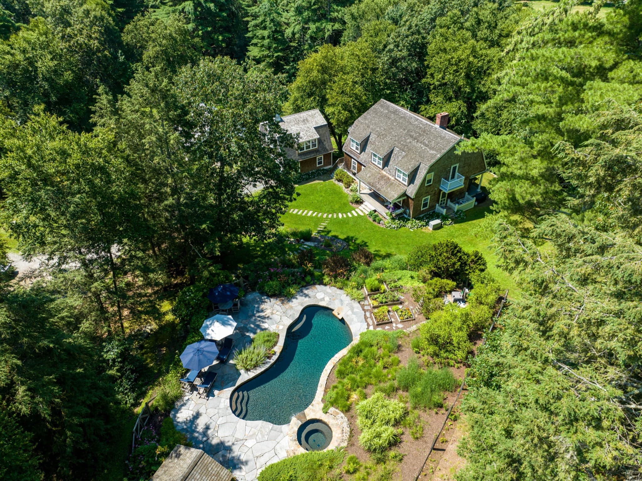 Aerial view of a wooded property with a pool, stone terrace, and planted gardens