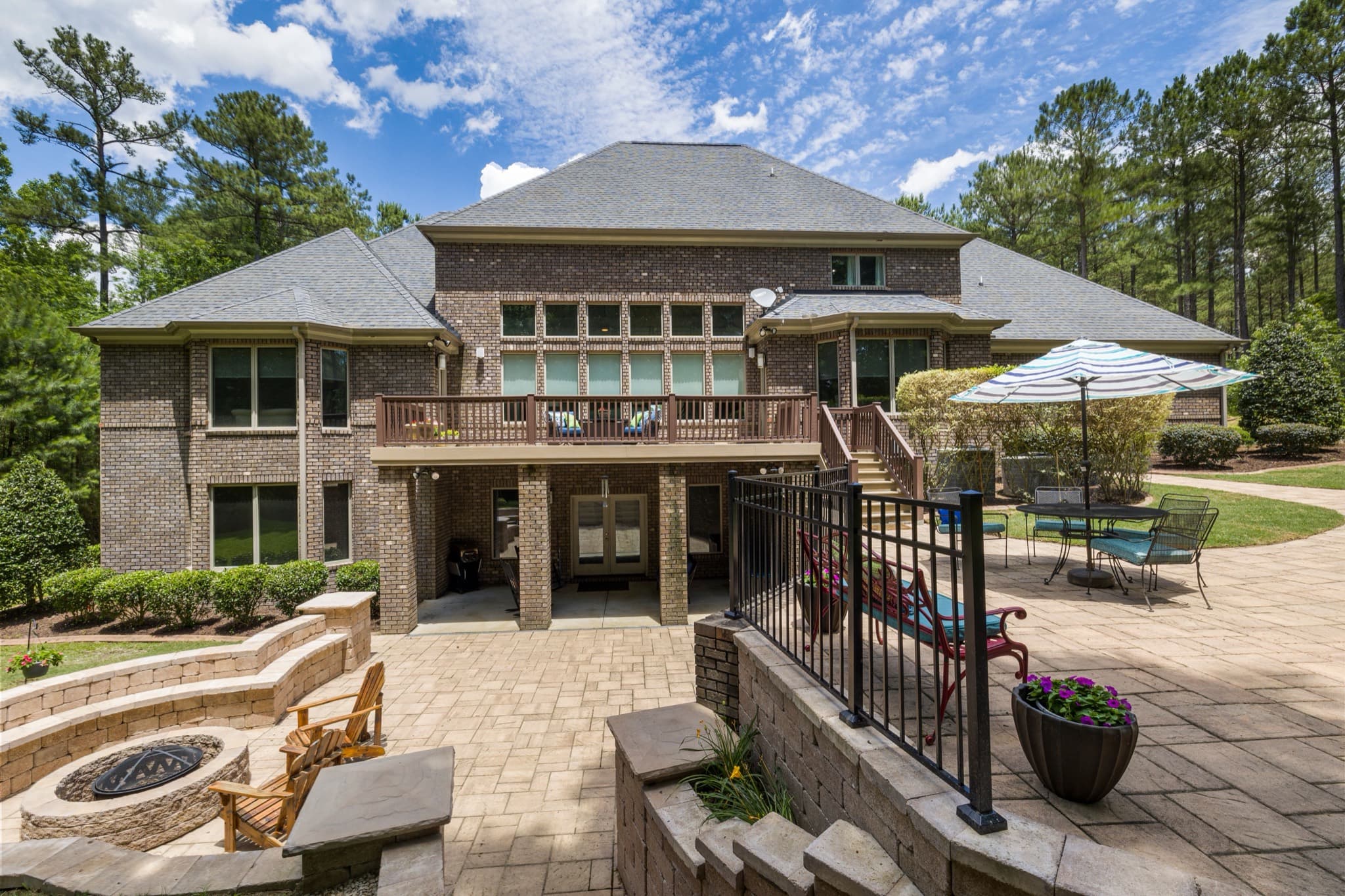 Back of a brick home with a deck, stone patio, fire pit, and tall pines